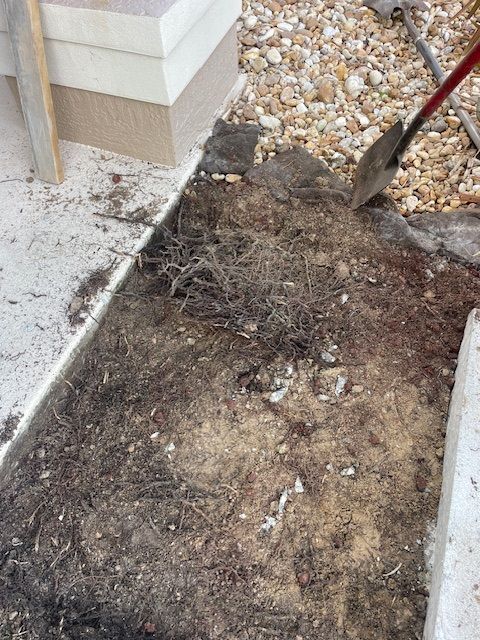A person uses a shovel to dig up dirt and weeds in a small garden bed next to a stone wall and walkway.