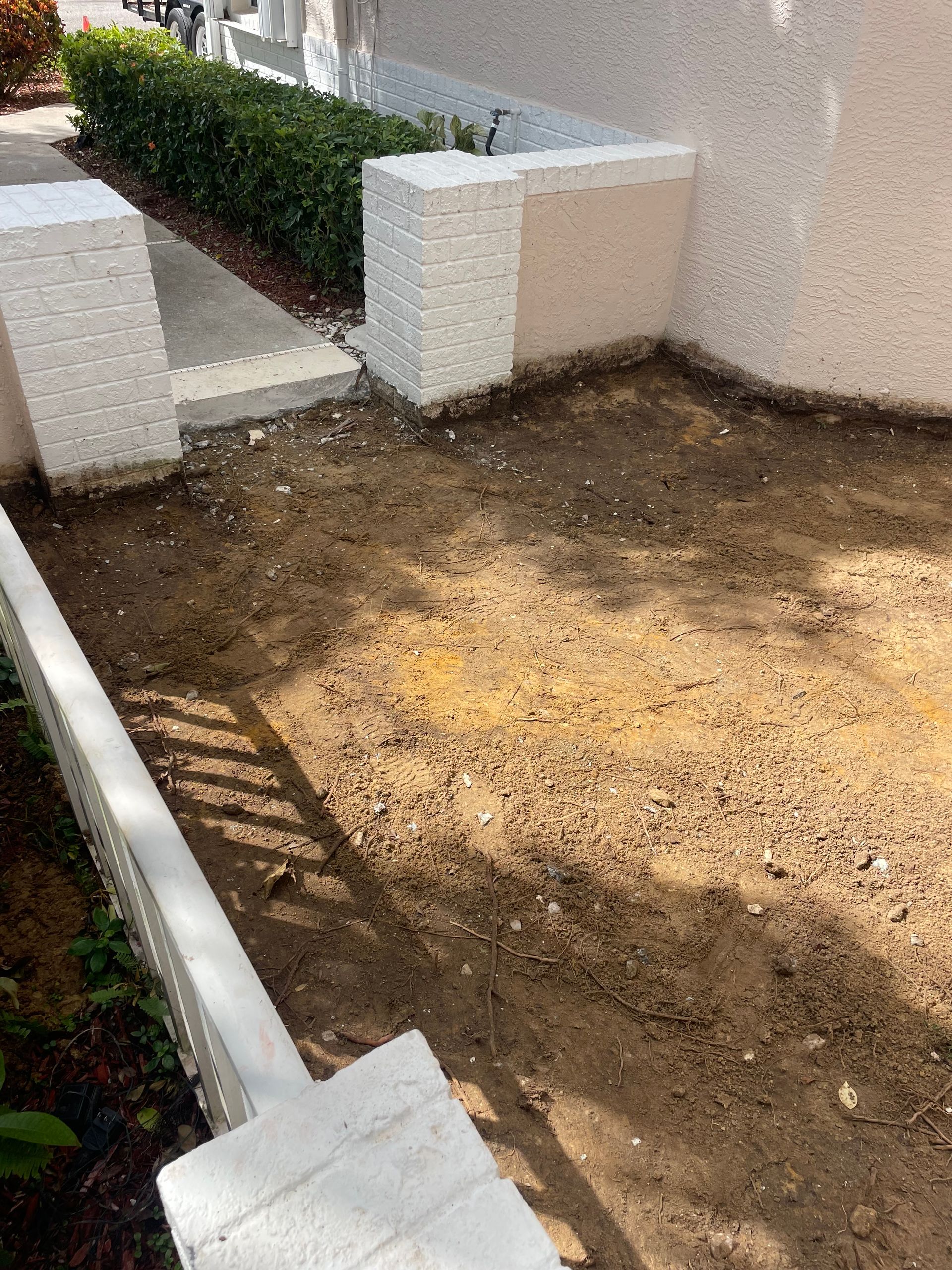 Dirt patch beside a white wall and low barrier; entrance path with green bushes in the background.