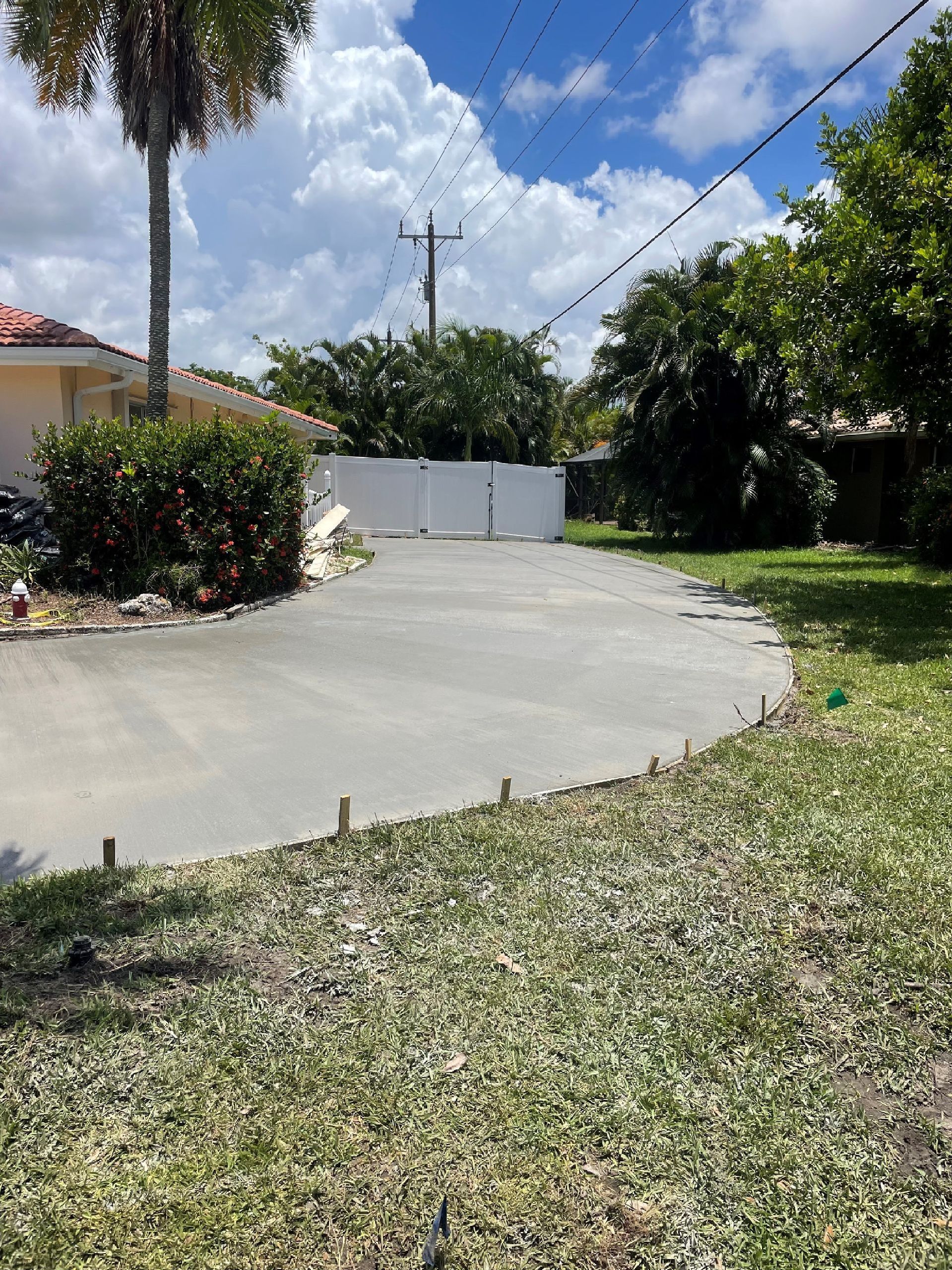 Newly poured concrete driveway, surrounded by grass, with a white wall at the end. Blue sky and trees in the background.