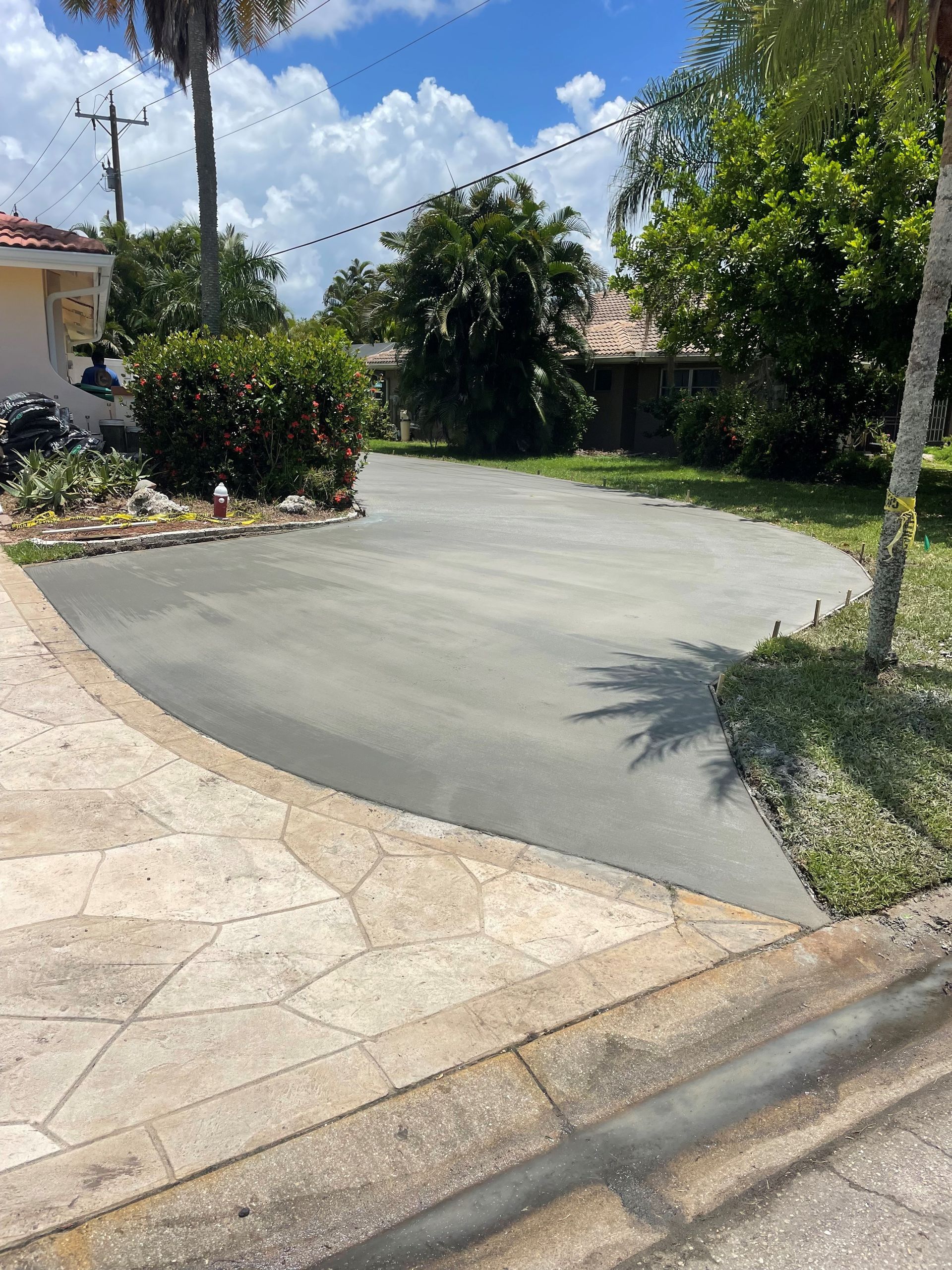 Asphalt driveway next to a patterned stone walkway and grass, with palm trees and a house in the background.