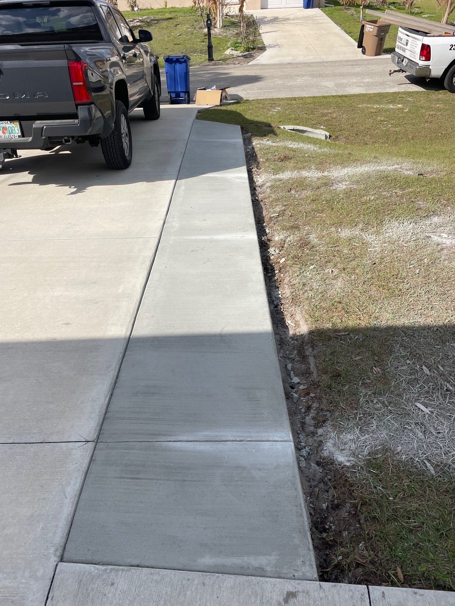 A newly poured concrete walkway extension runs parallel to a driveway, bordered by a grassy yard and a parked truck.