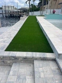 Green turf strip between gray stone walkways, beside water and buildings.
