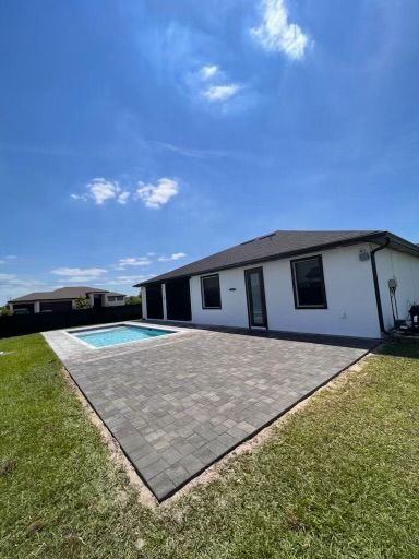 Backyard with patio, pool, and white house under blue sky.