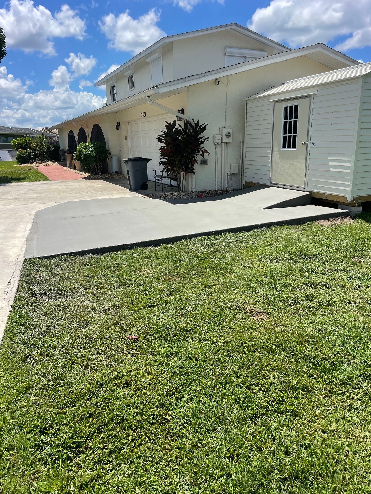 Side view of a two-story white house with a concrete patio and small shed in front of a grassy yard, blue sky above.