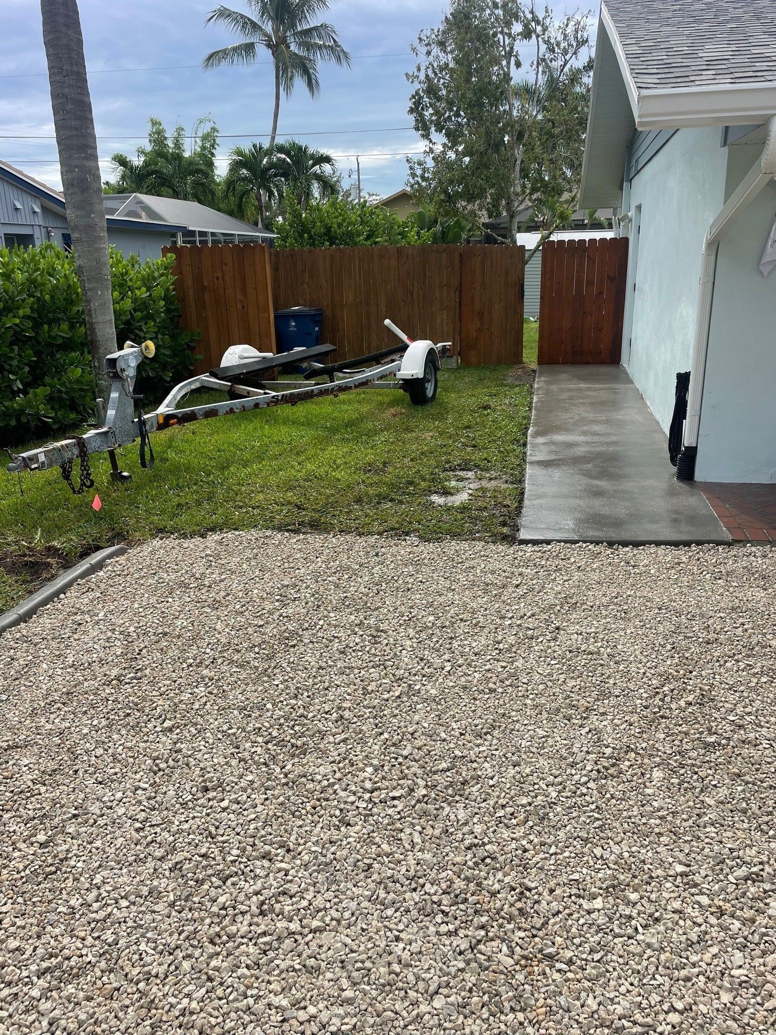 Gravel driveway beside a white house, leading to a small lawn with work materials and a wooden fence.