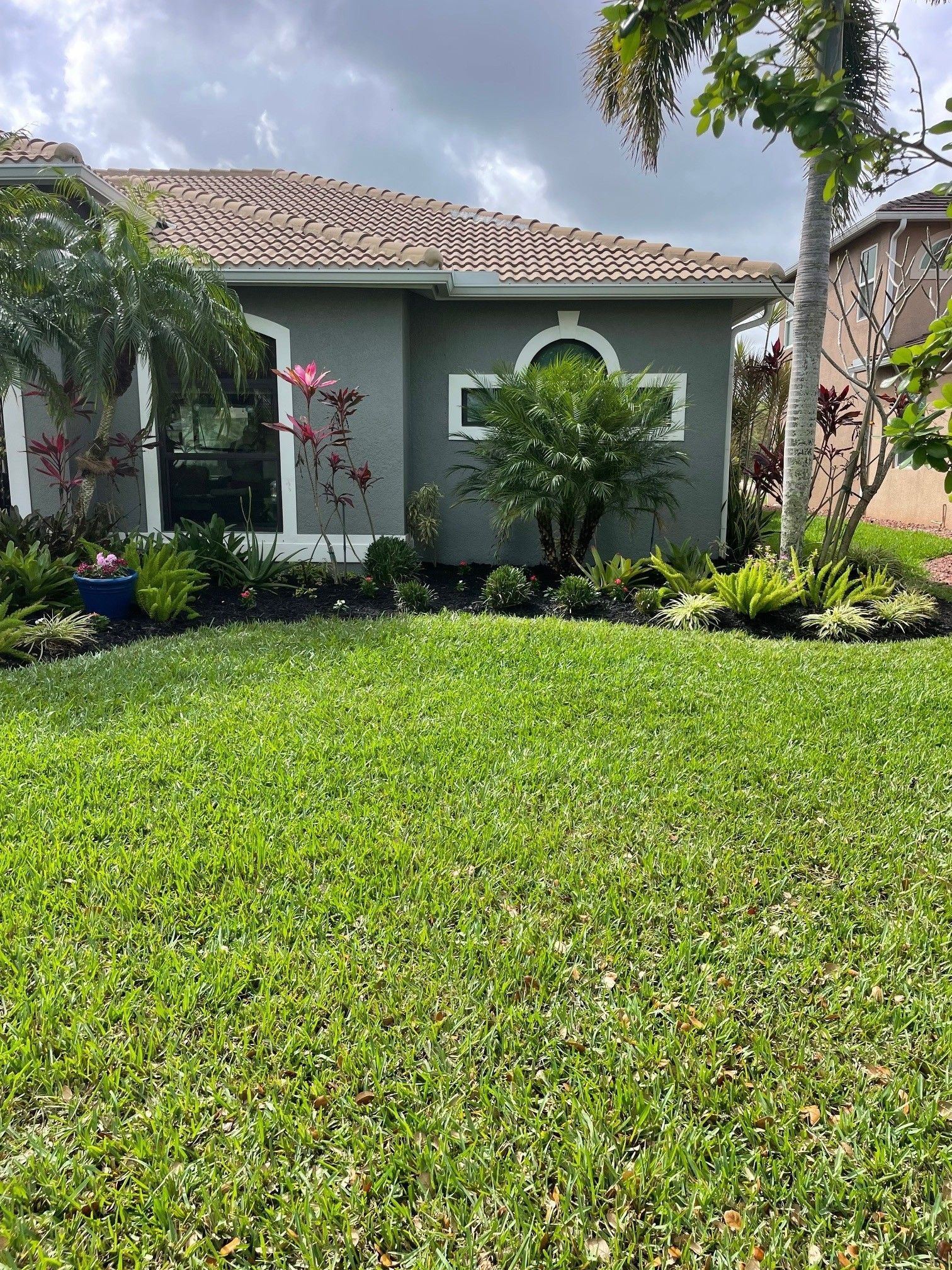 A grey house with a brown tiled roof, large front windows, and lush green landscaping against a cloudy blue sky.