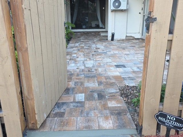 A stone-paved patio seen through an open wooden gate leading to a house exterior.