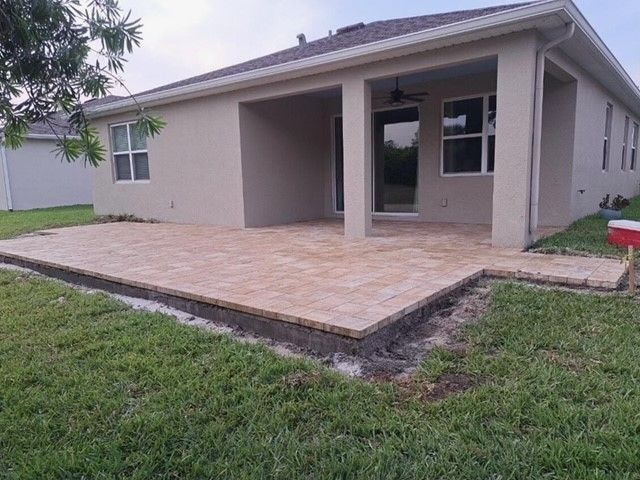 Backyard patio with light brown pavers, tan house, and green grass.