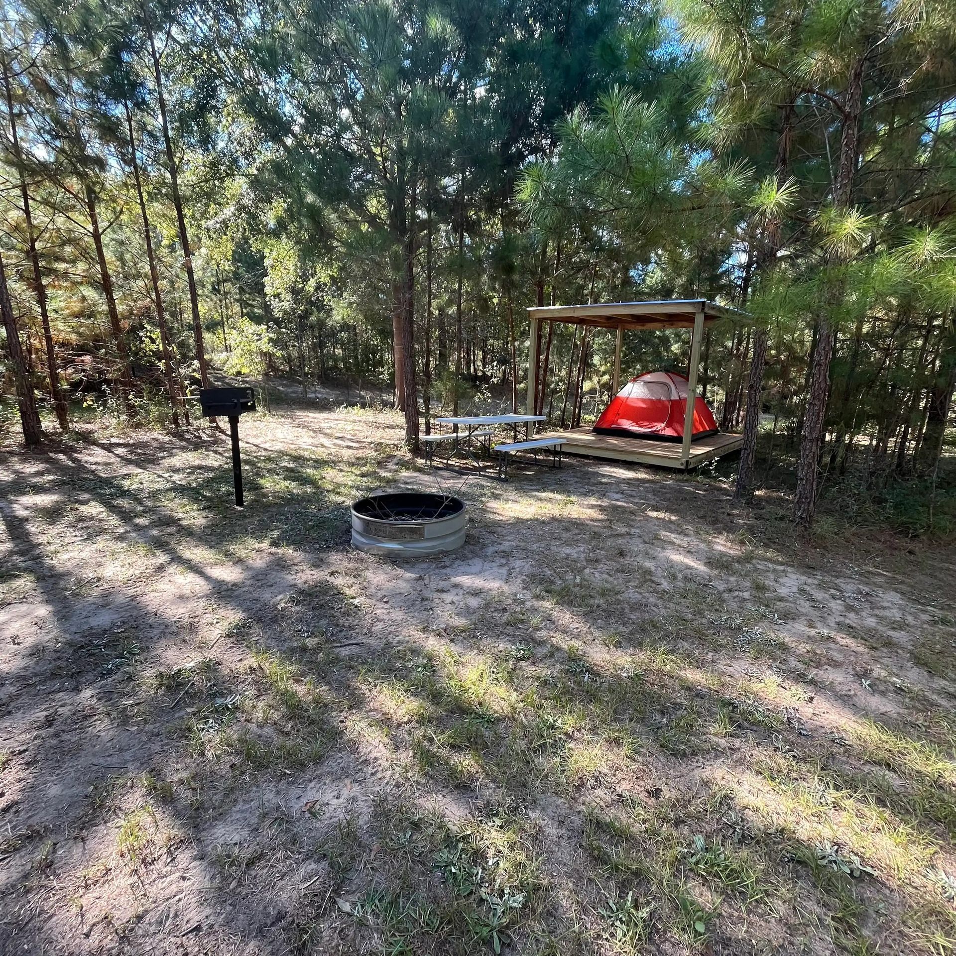 Wooded campsite with a picnic shelter, red tent, fire ring, and grill in dappled sunlight