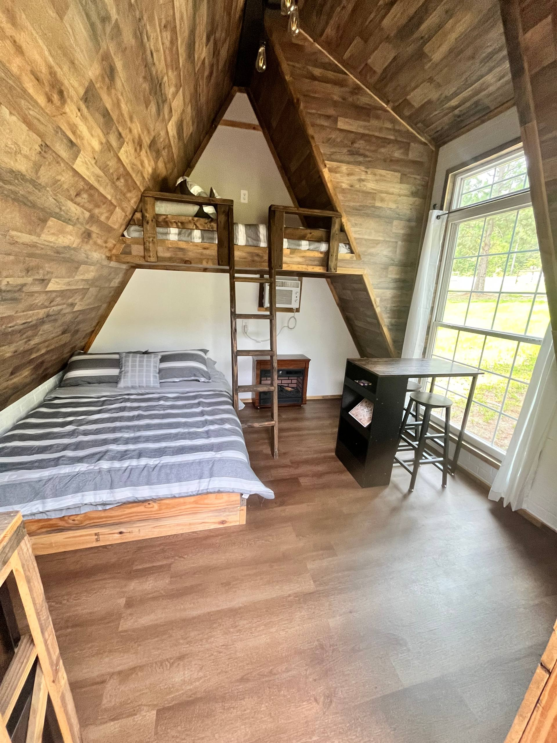 Cozy loft bedroom with a bed, desk, ladder shelf, and large window under a wooden sloped ceiling