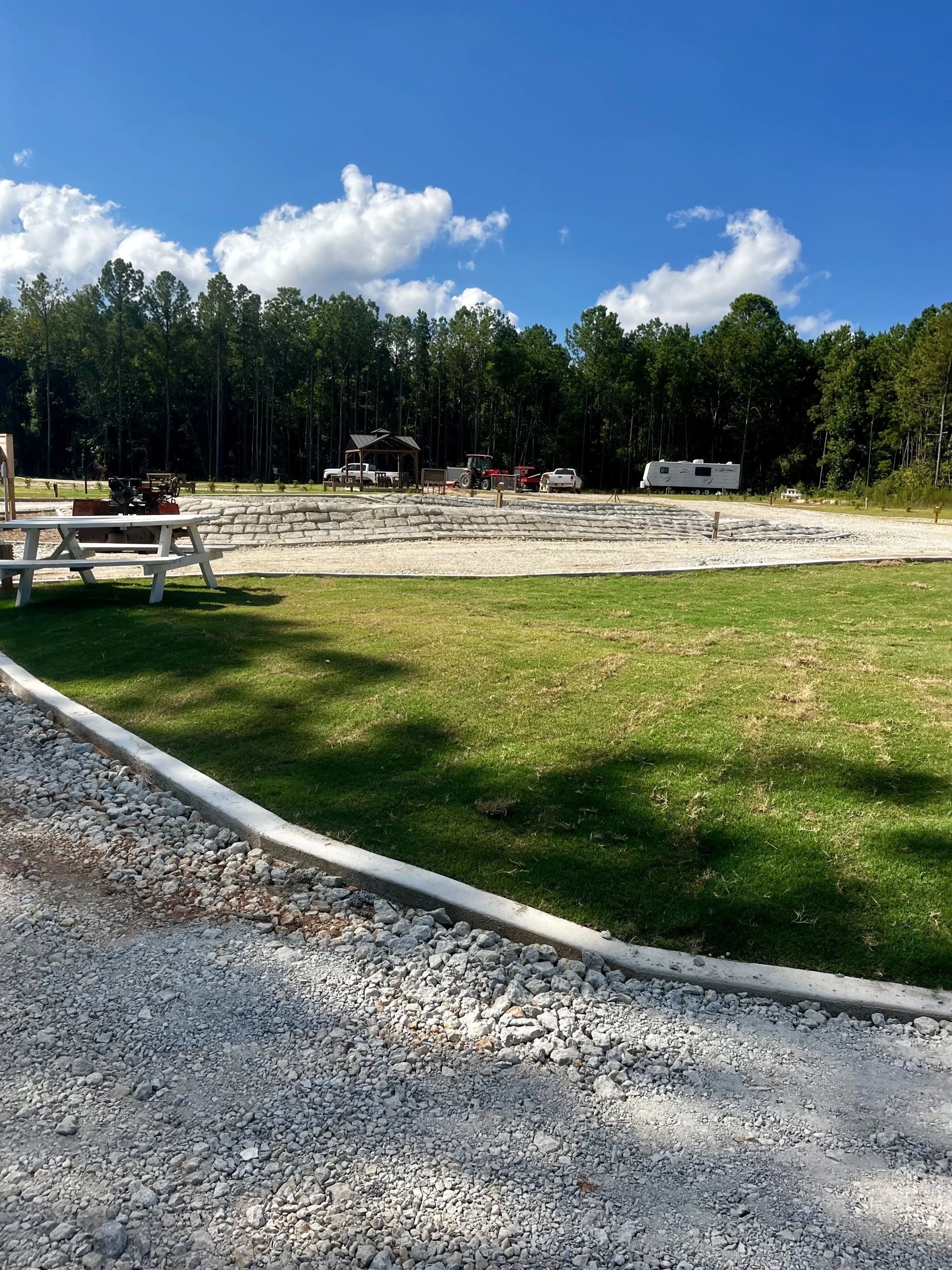 Gravel path beside a grassy picnic area with benches, trees, and a blue sky with clouds