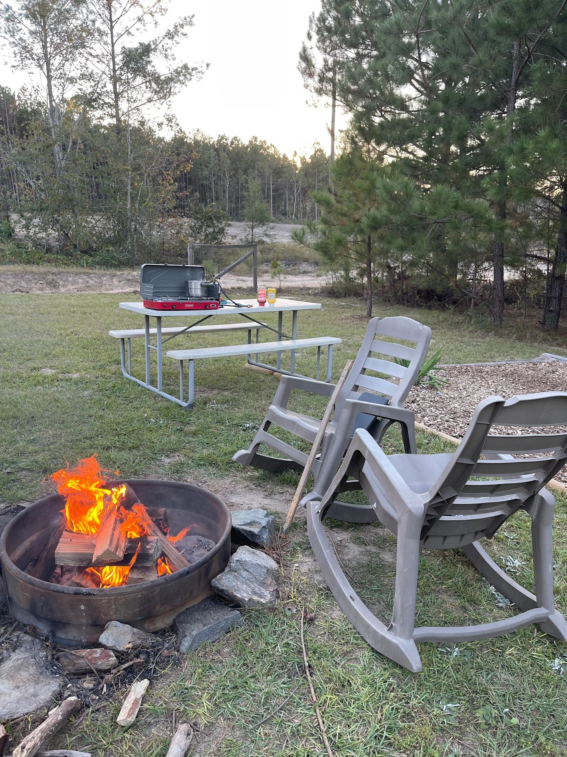 Fire pit with chairs in a grassy campsite, with a table and grill nearby under trees at dusk