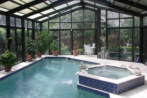 Indoor pool and hot tub in a glass-walled room, with potted plants and a view of greenery outside.