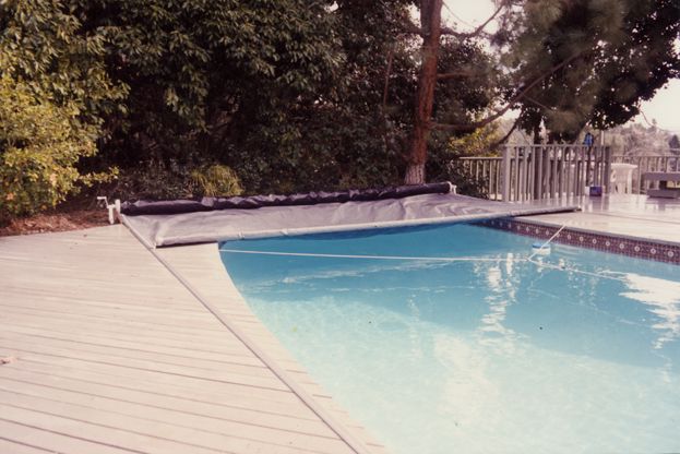 Backyard swimming pool beside a wooden deck, surrounded by trees and a fence.