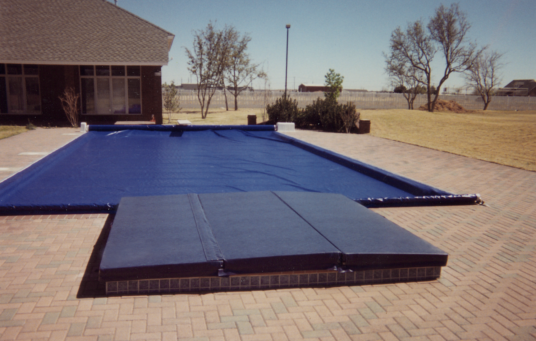 Covered backyard swimming pool with blue tarp and brick patio beside a house