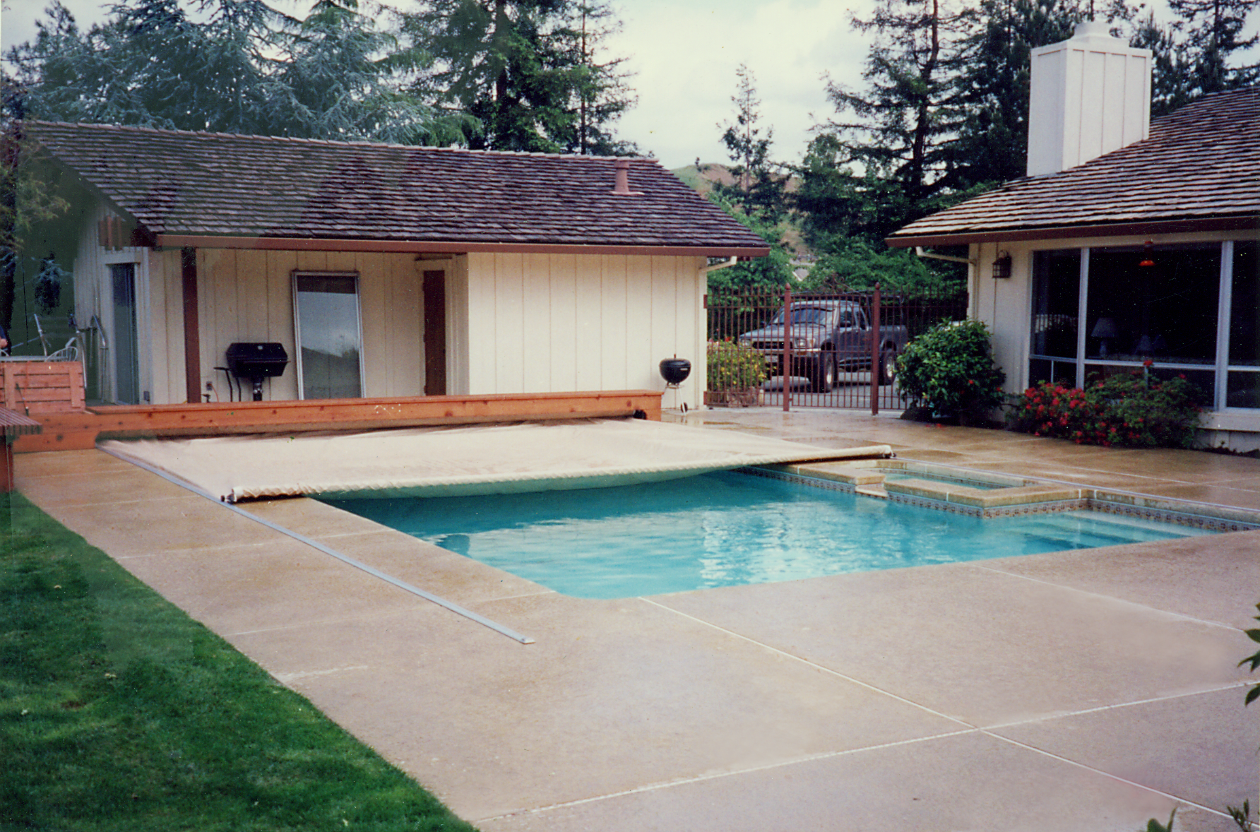 Backyard patio with a small swimming pool, concrete deck, and a shed-like pool house surrounded by trees.