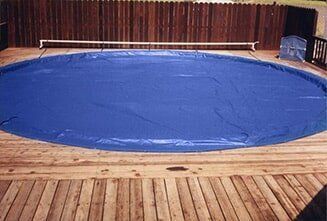 Large blue tarp or pool cover stretched over a circular area on a wooden deck indoors