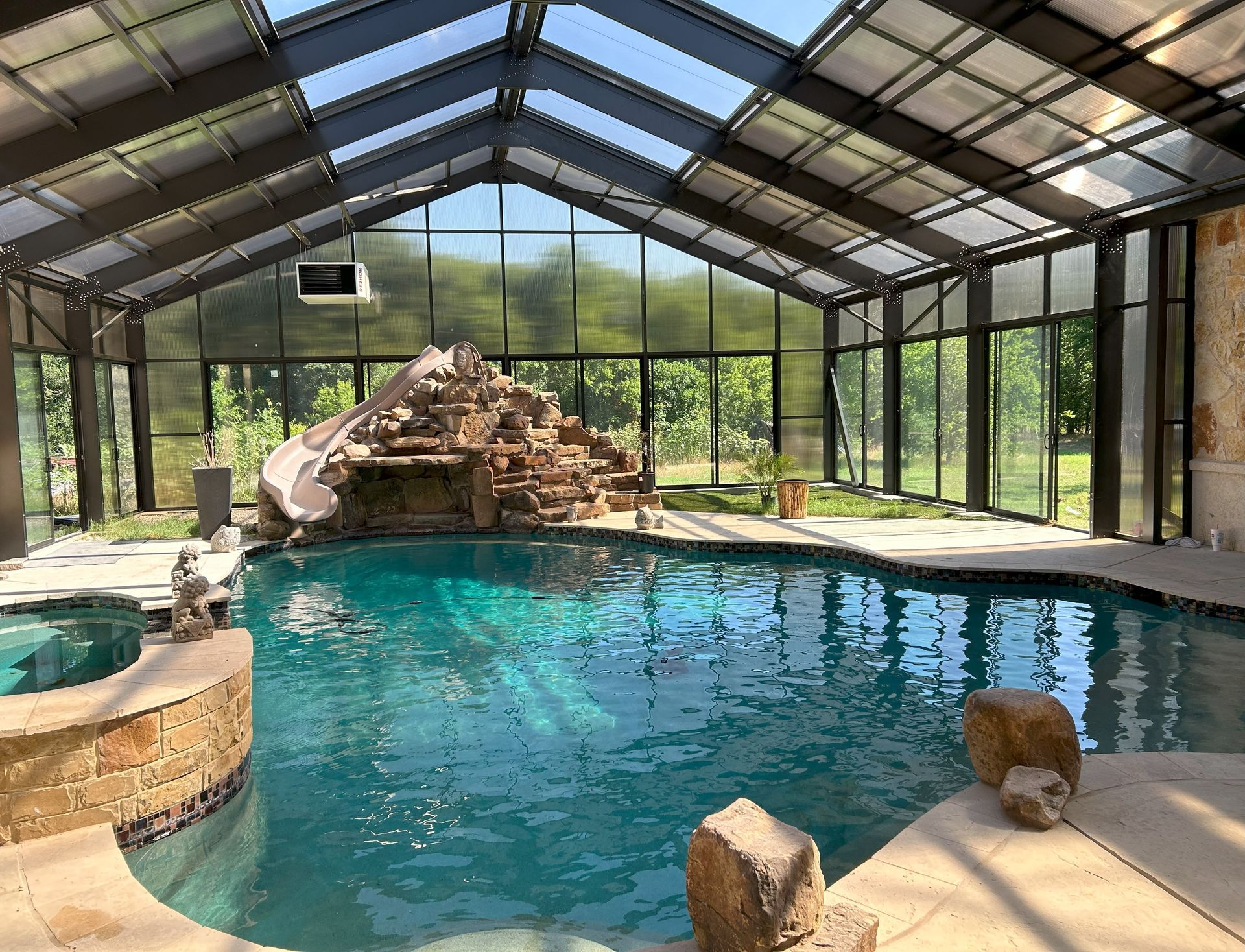Indoor pool with turquoise water under a glass roof, featuring a rock waterfall and a small animal on the deck