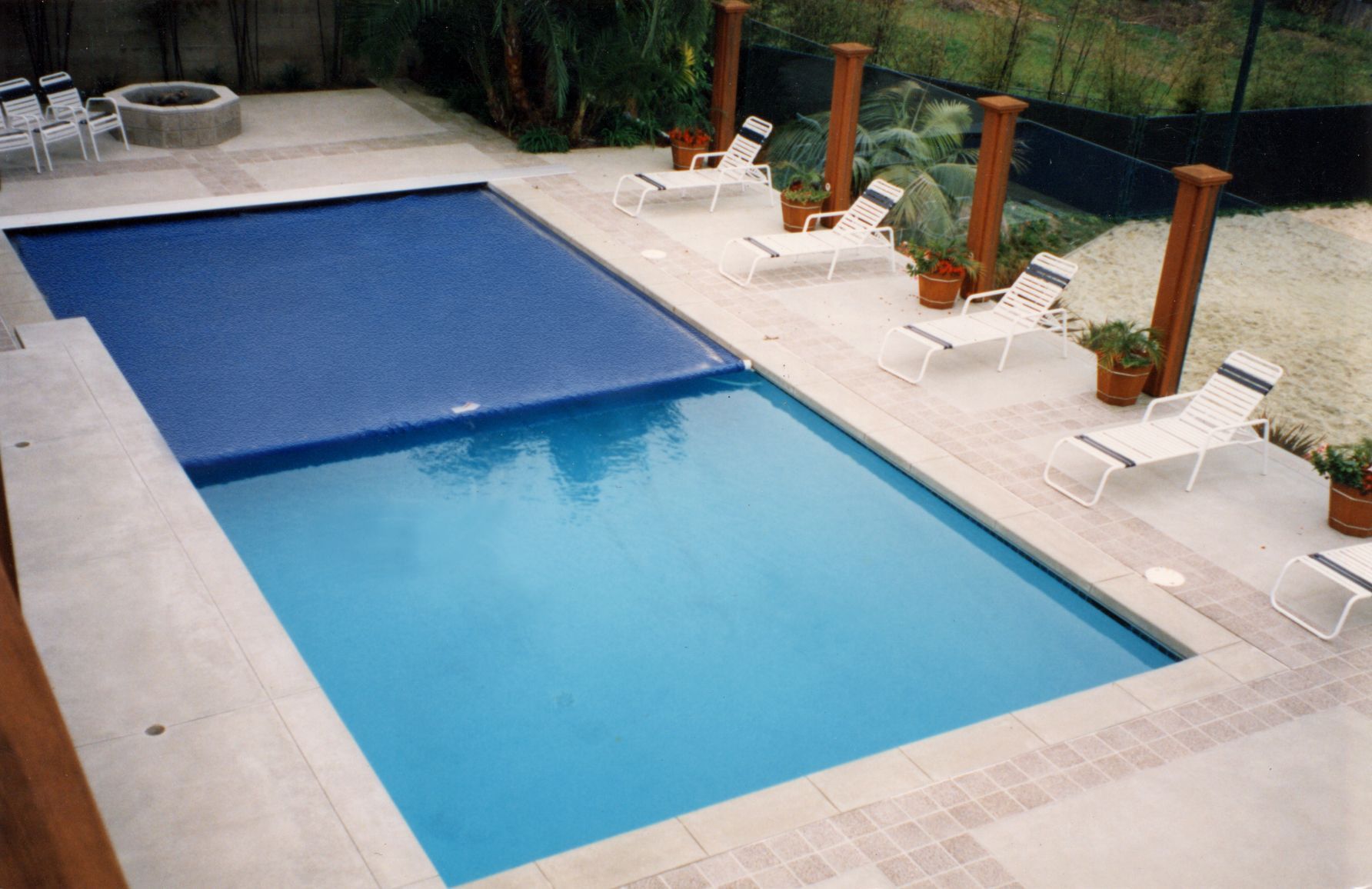 Outdoor swimming pool with a blue cover partially rolled back, beside white lounge chairs and potted plants
