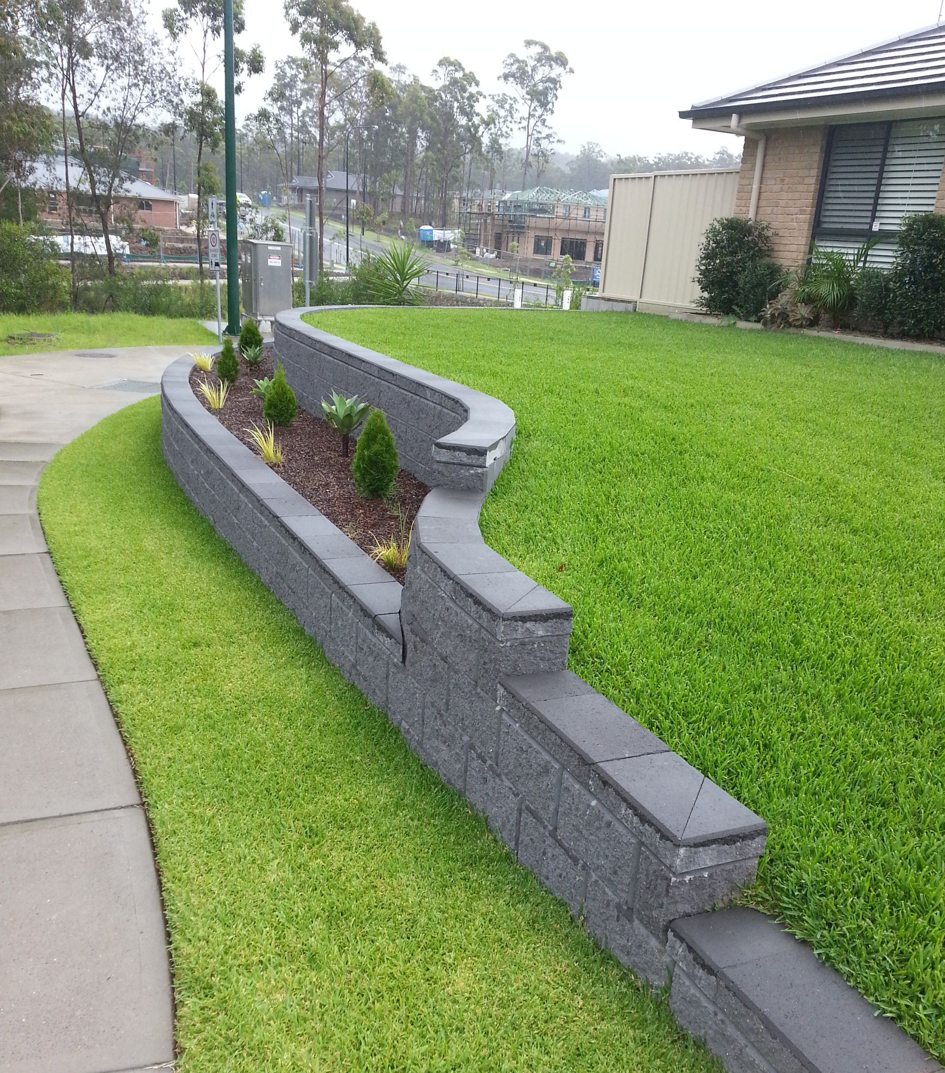 A gray stone retaining wall curves along a green lawn, with plants in a raised bed.