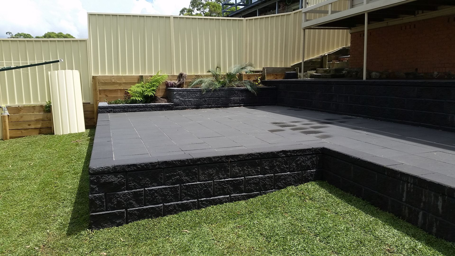 Black brick patio with grass lawn, beige fence, and green foliage.