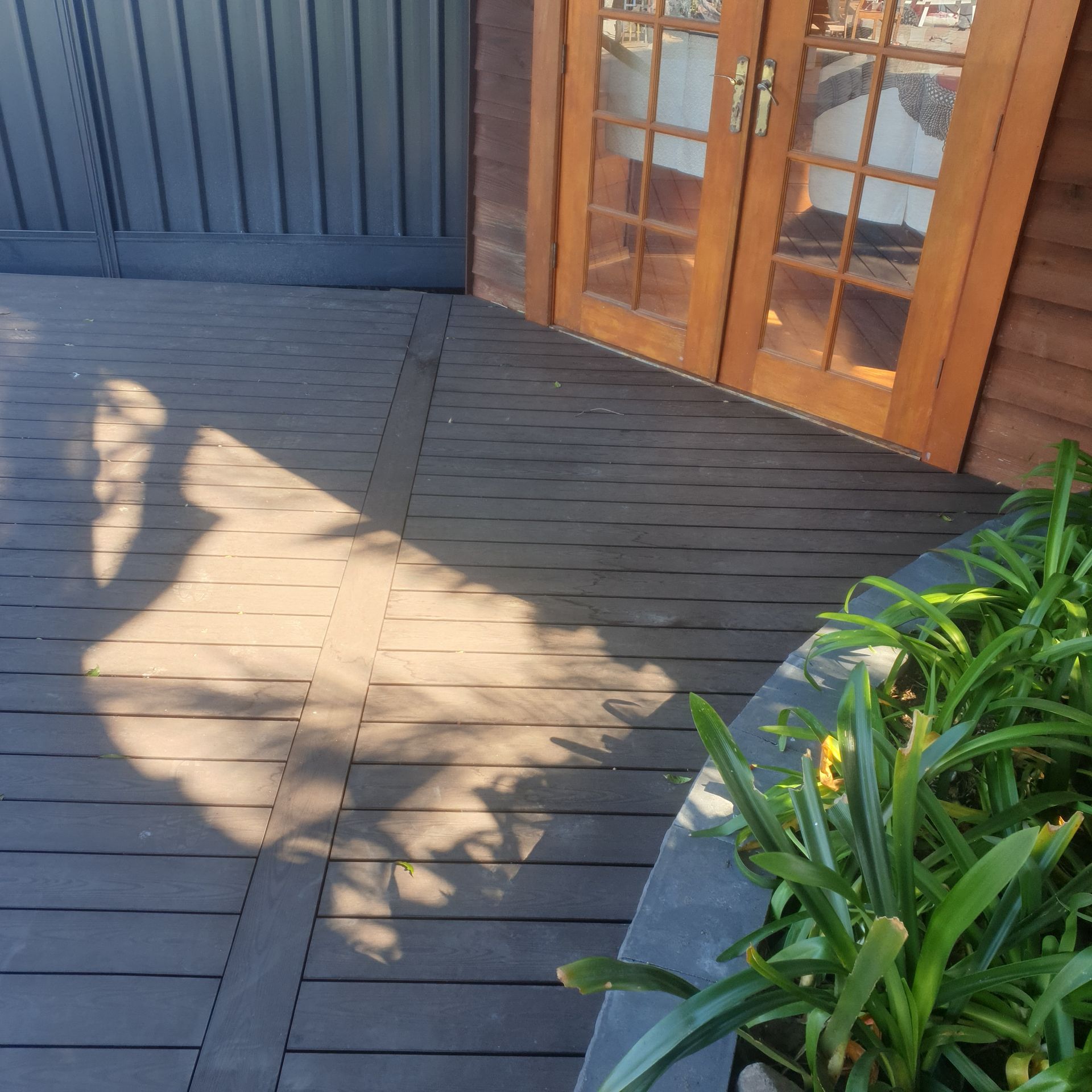 Wooden deck with shadow, next to wood door and plants.
