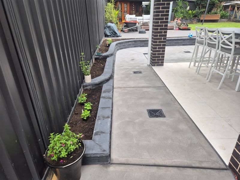 Cement walkway alongside a brick wall and fence. Brown dirt and green plants border the path.