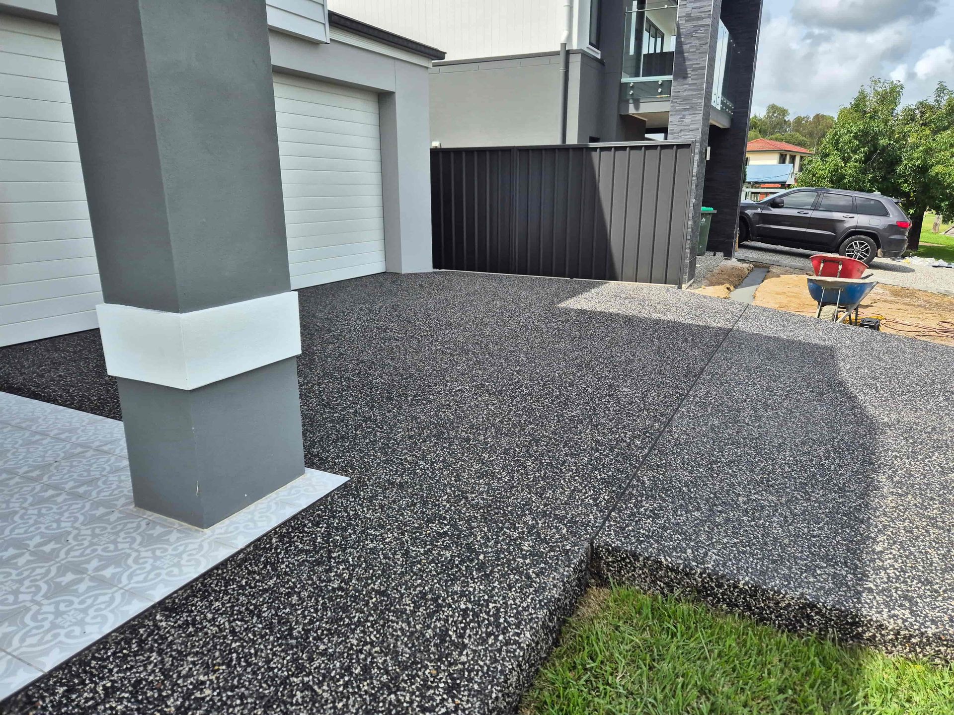Black speckled driveway with gray column, garage, and house. Green grass on the right.