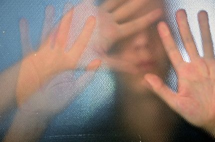 Mani premute contro una superficie traslucida e ruvida, con volti sfocati e una camicia blu sullo sfondo.