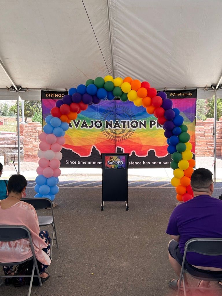 A group of people are sitting under a tent with a rainbow balloon arch.
