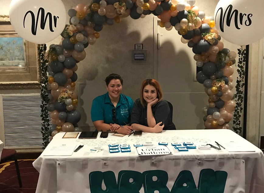 Two women are sitting at a table under a balloon arch.