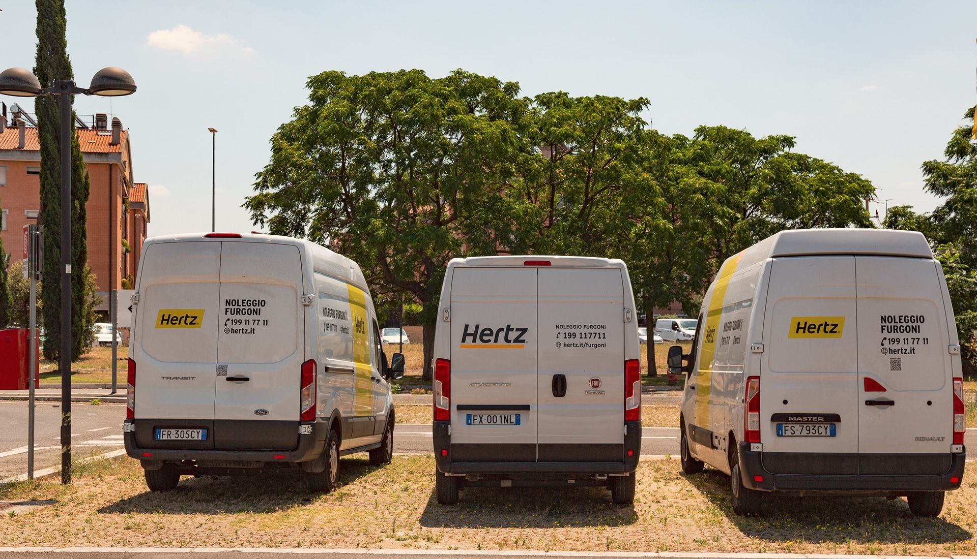 Three white Hertz rental vans parked in a gravel lot on a sunny day.