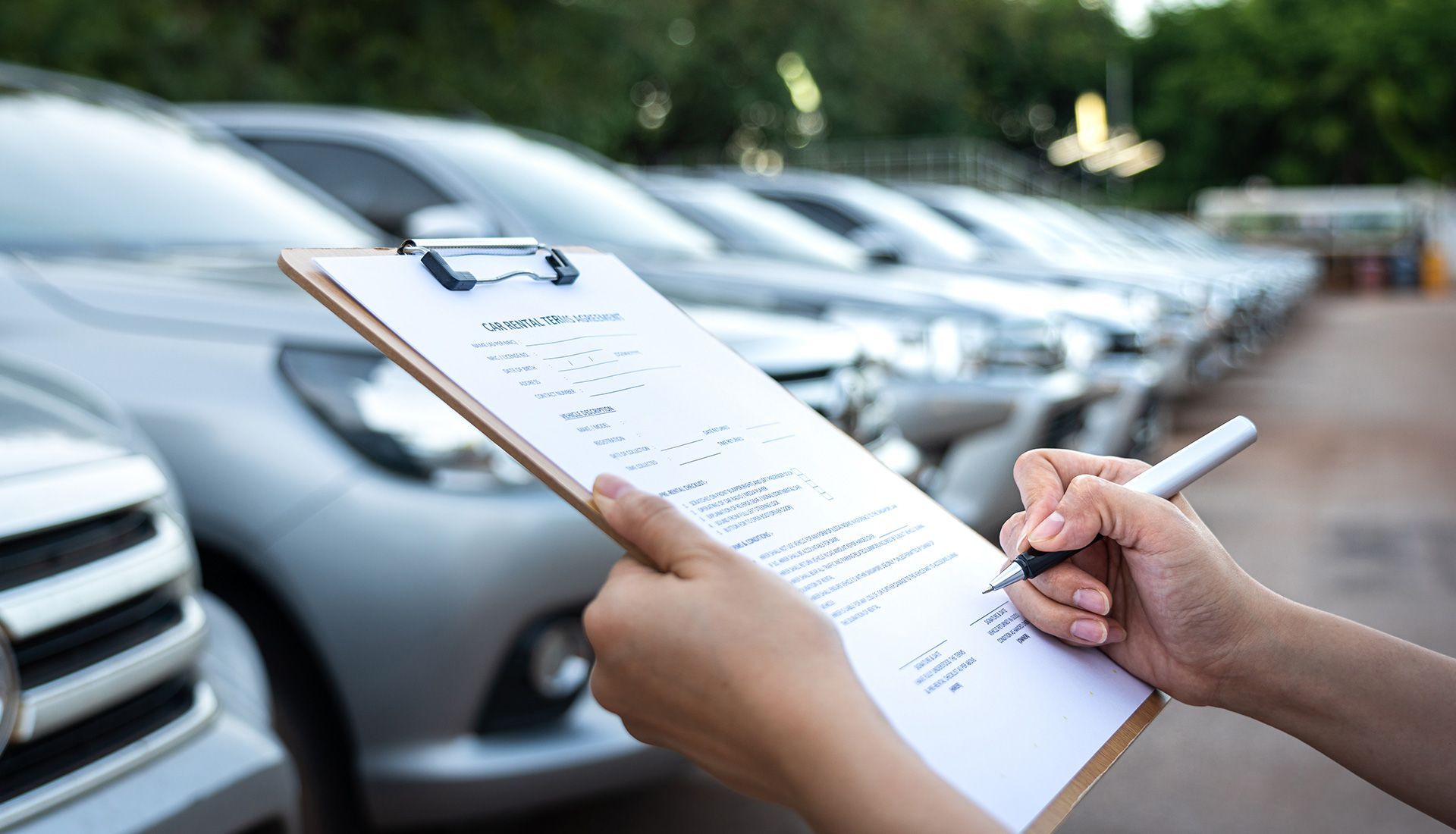 A person writes on a clipboard, inspecting silver cars parked in a lot.