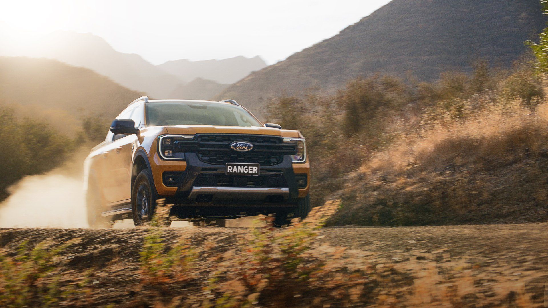 Tan Ford Ranger pickup truck driving on a dirt road, kicking up dust in a mountain landscape.
