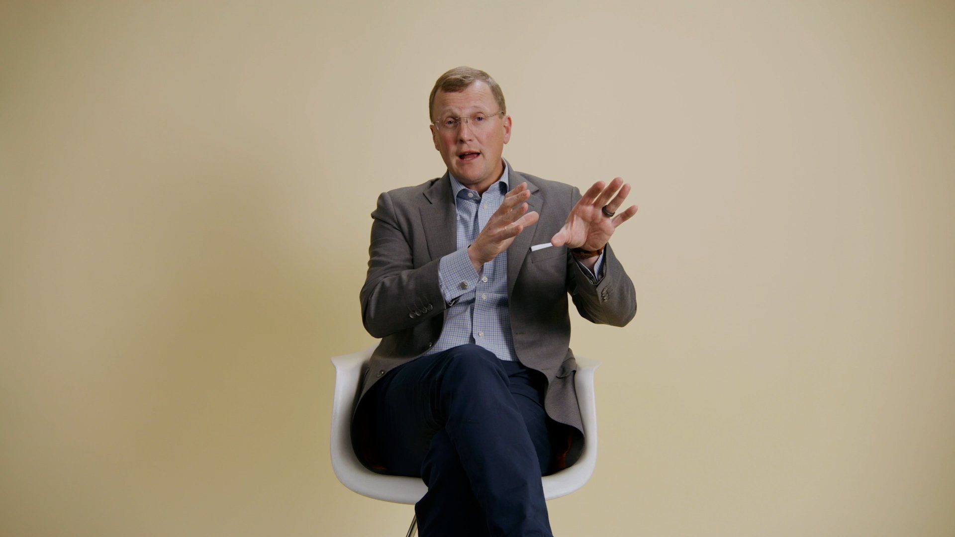 Man seated, gesturing with hands, wearing a blazer, light blue shirt, against a pale yellow backdrop.
