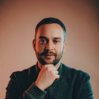 Man with a beard, holding his chin, looking at the camera. Warm lighting and a neutral background.