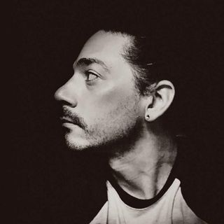 Man with slicked-back hair, looking left in profile, dark background, wearing a light-colored shirt.