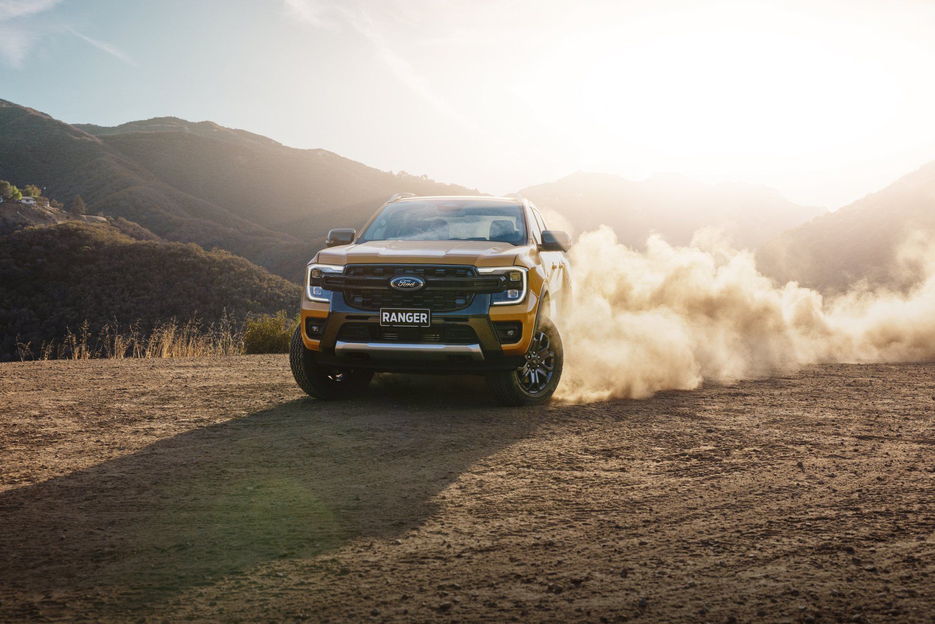 Yellow SUV kicking up dust on a dirt road in a mountainous, sunny landscape.