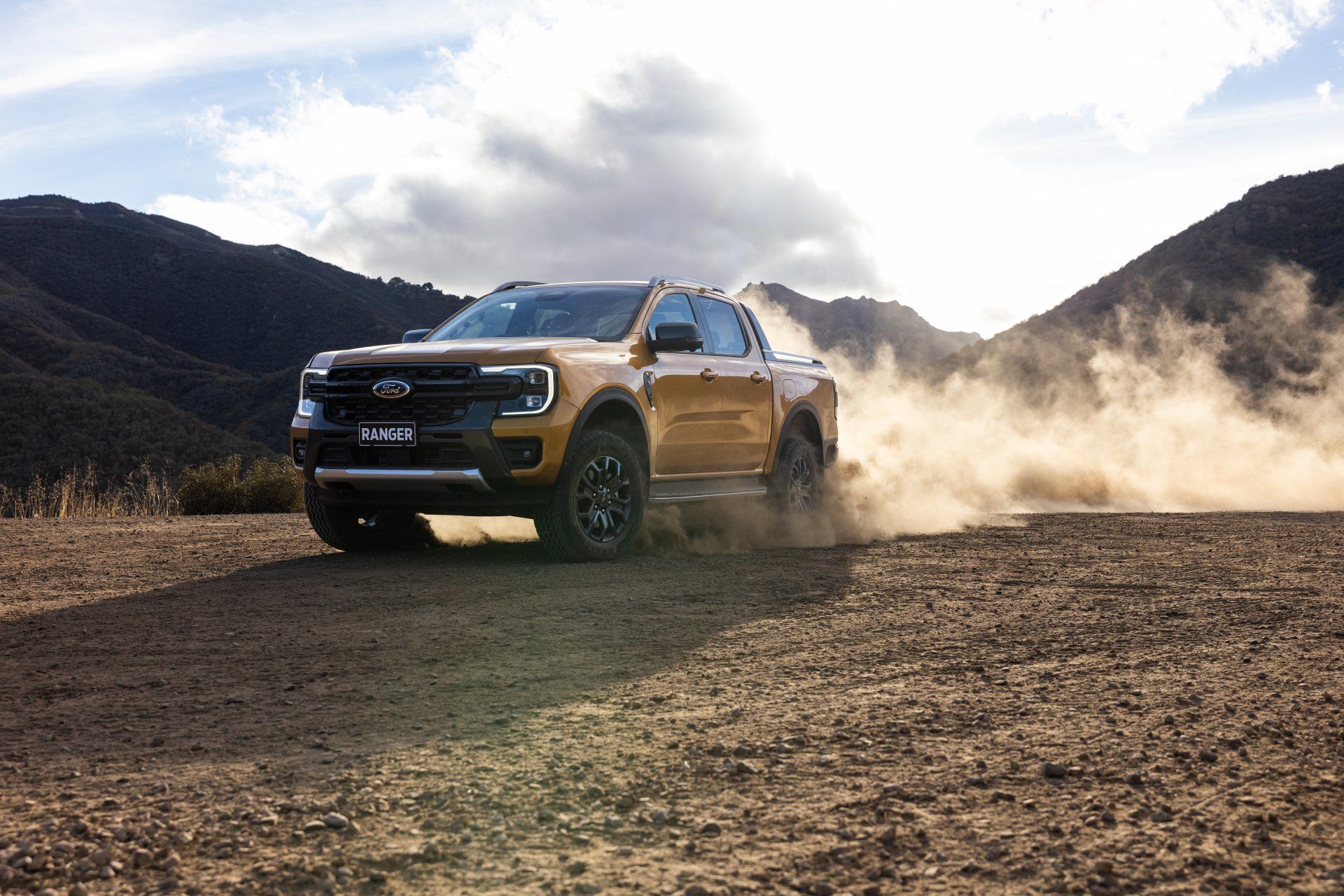 Yellow Ford Ranger truck kicking up dust on a dirt road in a mountainous area.