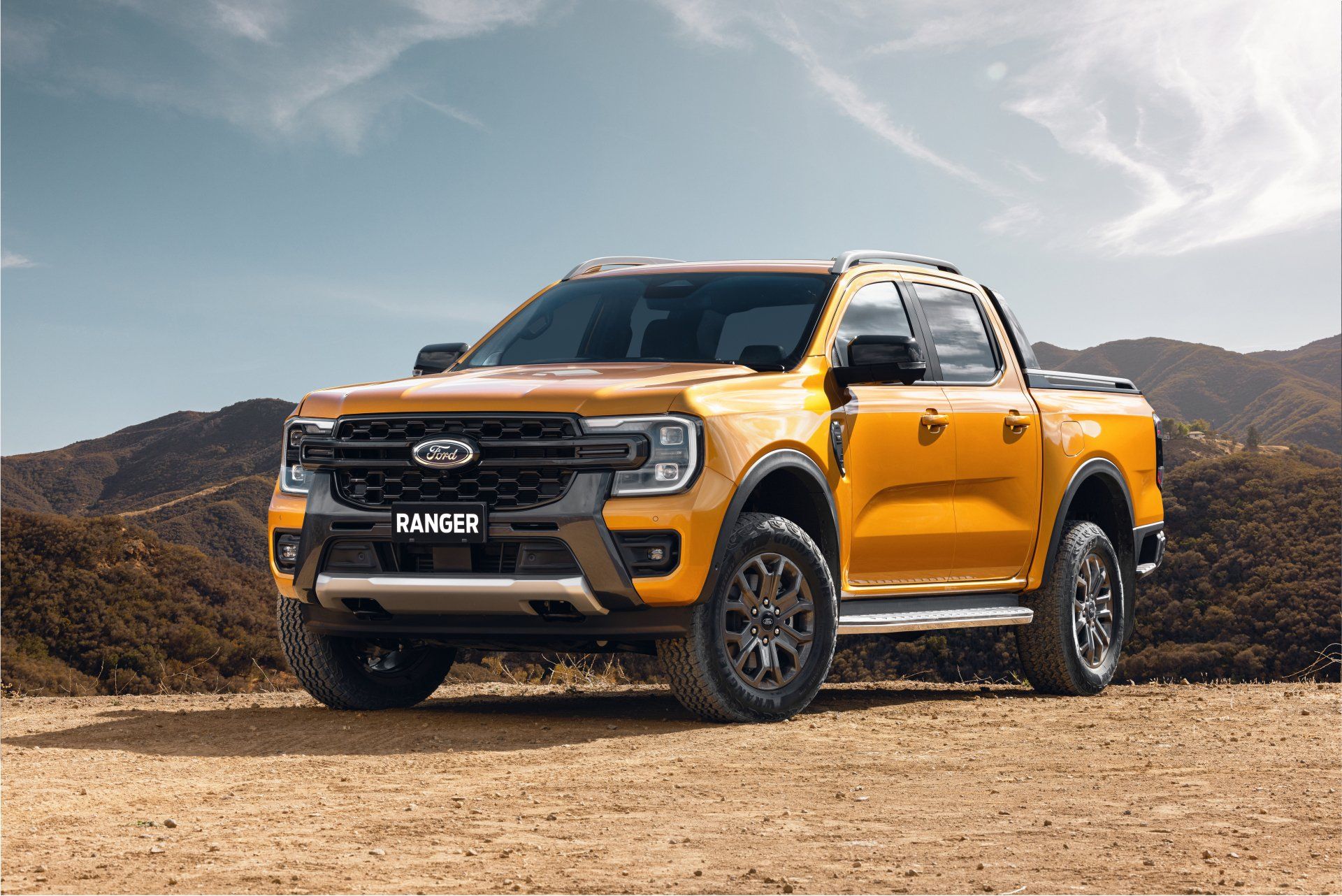Yellow Ford Ranger pickup truck parked on dirt road in front of hills under a blue sky.