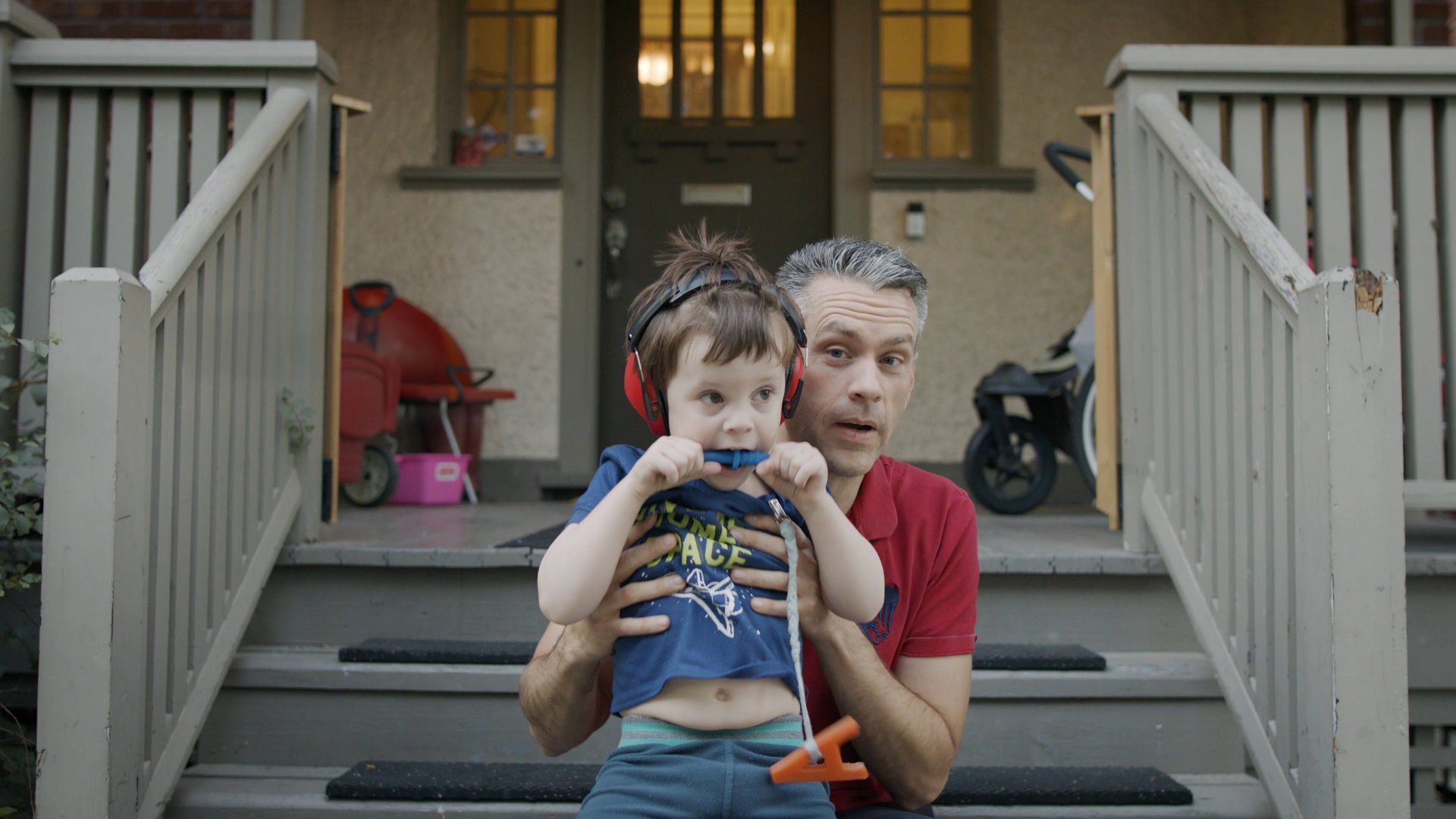 Man holding a child wearing ear protection and pacifier on porch steps. Red shirt.