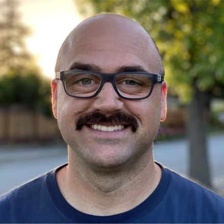 Bald man with glasses and mustache smiling outdoors.