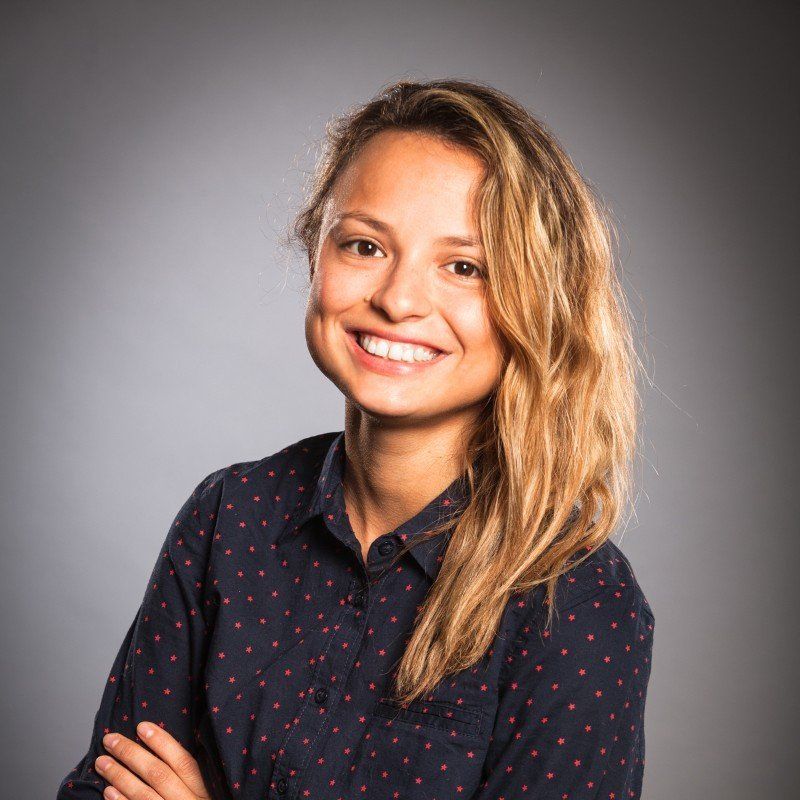 Woman with long, wavy blonde hair smiles, arms crossed, wearing a navy shirt with red dots, against a gray background.