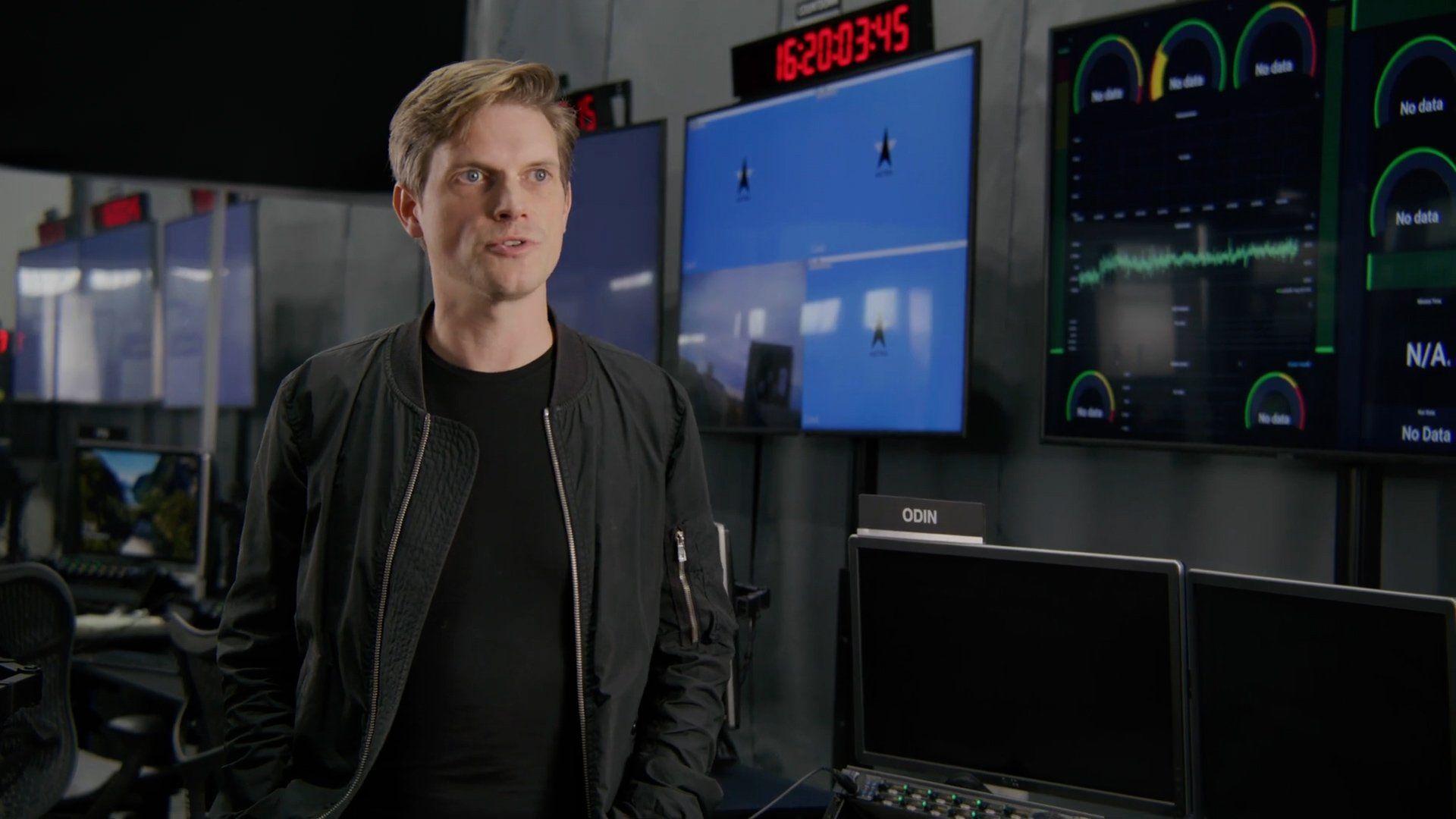 Man in a black jacket speaking in a control room with monitors displaying data and time.