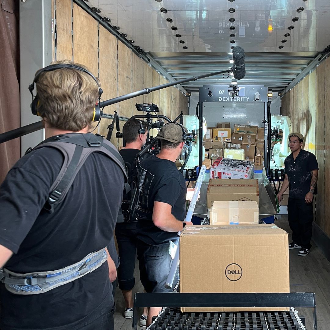 Film crew inside a truck. Crew members with equipment surround boxes. Interior truck filled with more boxes.