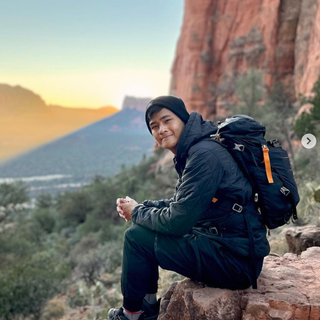 Man sitting on a rock overlooking a mountain range, wearing a backpack and winter clothing.