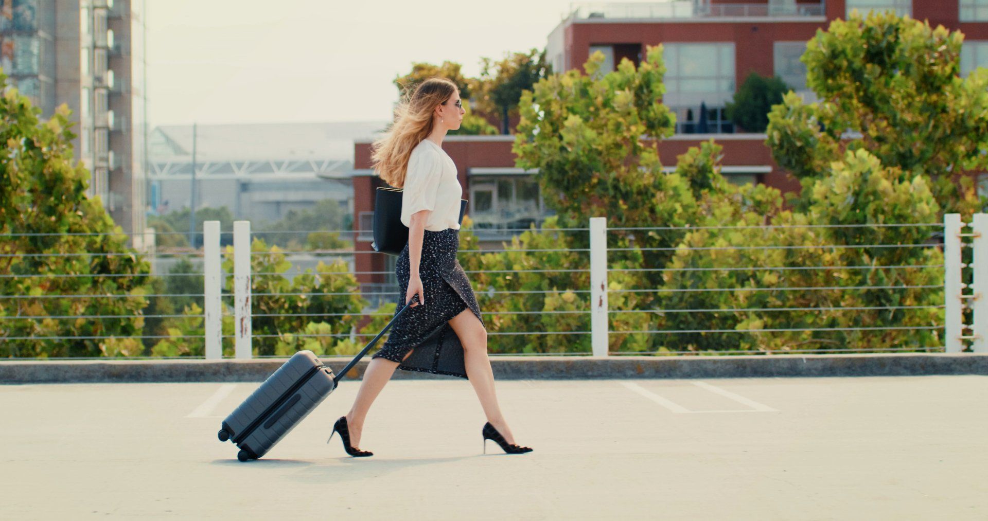 Woman with suitcase walking outdoors, wearing a skirt, heels, and backpack.
