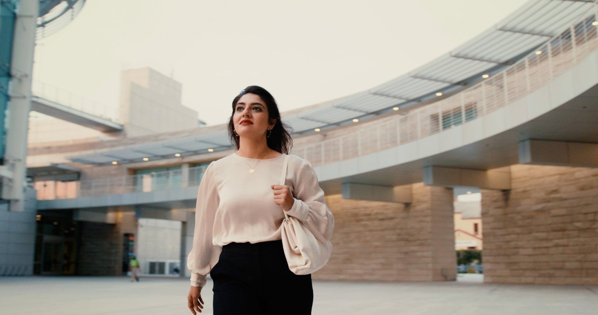 Woman in white top and black pants walks outdoors, carrying a purse, near a modern building.
