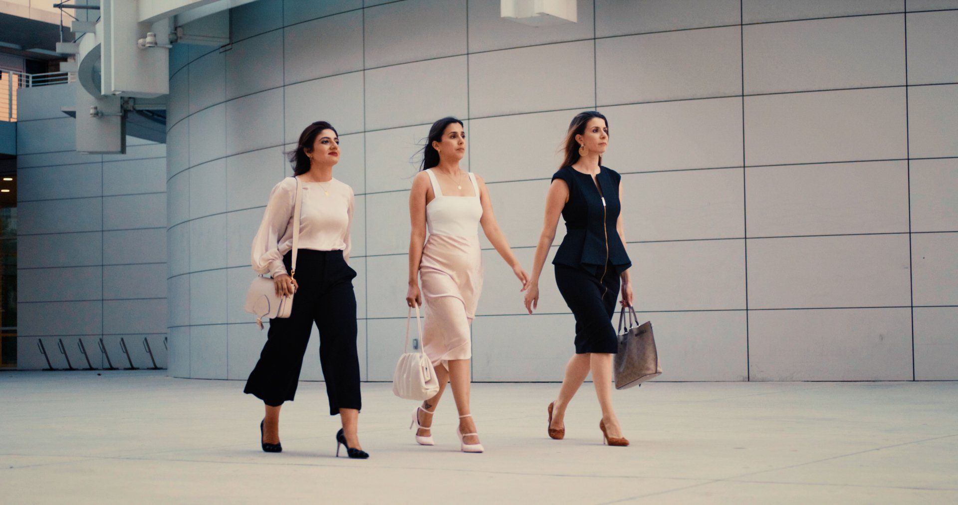 Three women walk outdoors, holding hands. Modern architecture background.