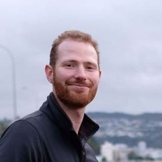 Man with red hair and beard smiling outdoors, wearing a black jacket; blurred cityscape background.